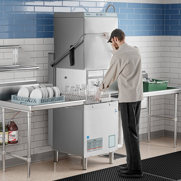 A man in a white coat and gloves putting glasses in a Noble Warewashing door-type dishwasher on a counter in a school kitchen.