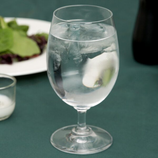 A close-up of a Stolzle water goblet filled with ice water on a table with a plate of salad.