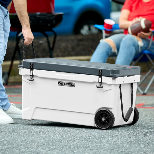 A person wheeling a white CaterGator outdoor cooler with black wheels.