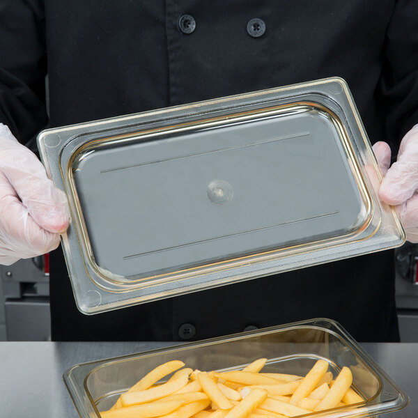 A person in gloves holding a Cambro plastic container lid over a tray of food.