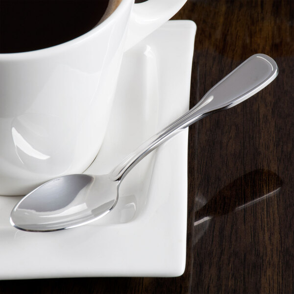 A Oneida Stanford stainless steel demitasse spoon on a saucer next to a cup of coffee.