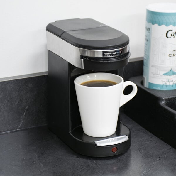 A Hamilton Beach stainless steel single serving pod coffee maker on a counter with a cup of coffee.