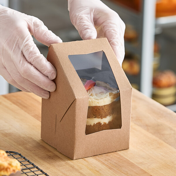A person in gloves holding a Baker's Lane brown Kraft window box with a piece of cake inside.