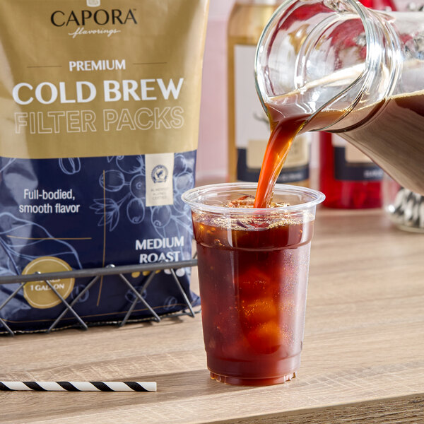 A plastic cup filled with ice is being poured with cold brew coffee from a glass pitcher, with a bag labeled 'Cold Brew Filter Packs' in the background.