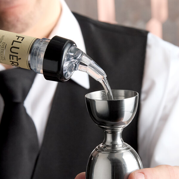 A close-up of a bottle with a measured liquor pourer dispensing liquid into a stainless steel jigger.