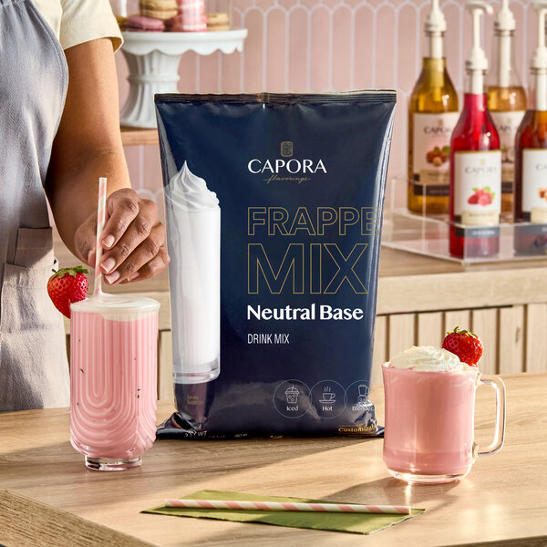A 3.5 lb. bag of Capora Neutral Base Frappe Mix displayed on a counter with two pink strawberry beverages.