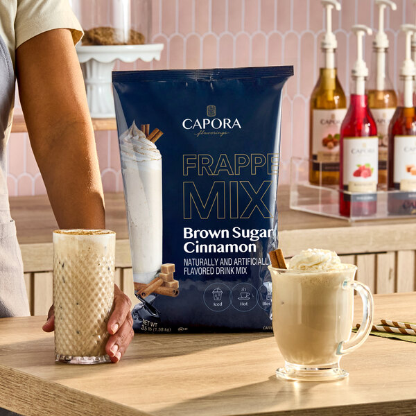 A bag of Capora Brown Sugar Cinnamon Frappe Mix weighing 3.5 lb is displayed on a counter with two prepared drinks beside it.