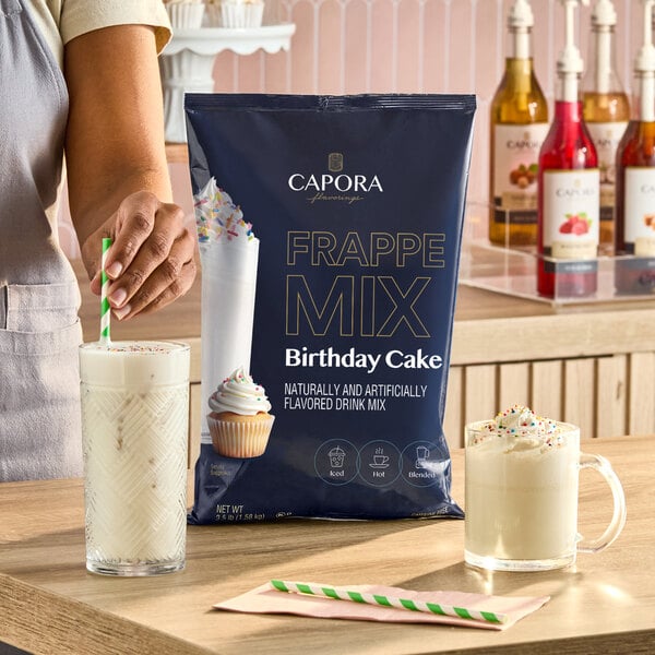 A bag of Capora Birthday Cake Frappe Mix displayed on a counter with two glasses of prepared birthday cake-flavored drinks.
