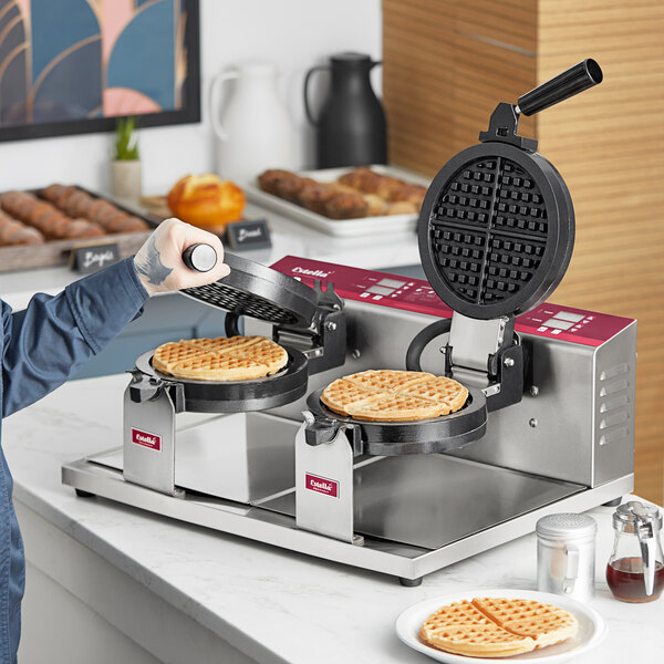 A double waffle maker with two round waffle plates, one open and one being closed, on a countertop with waffles inside.