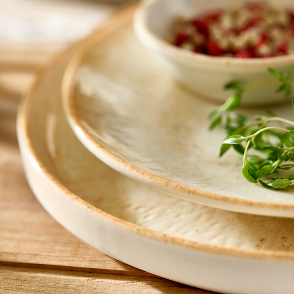A round beige porcelain plate with a slightly rustic edge, shown stacked with other plates and a small bowl.