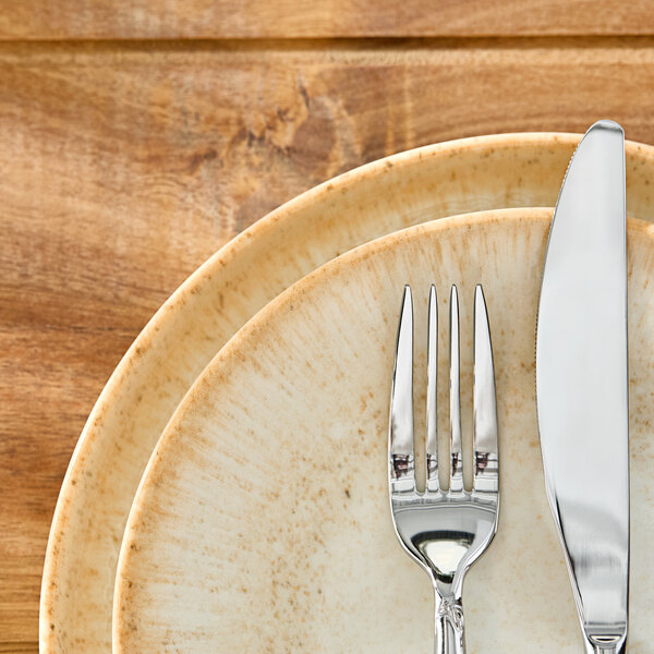 A round beige porcelain plate with a subtle speckled pattern, shown with a fork and knife on top.