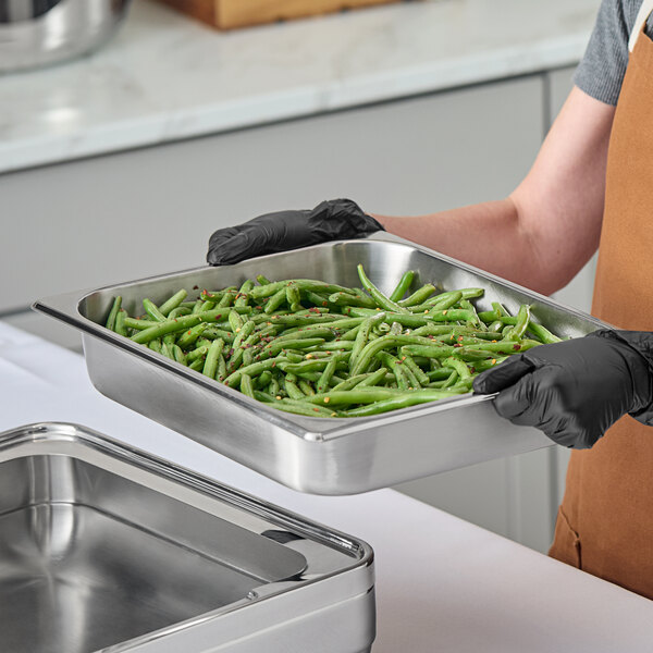 A stainless steel food pan filled with green beans being held by a person wearing black gloves.