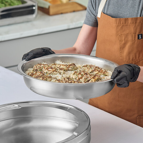 A round stainless steel induction chafer filled with food, being held by a person wearing black gloves and a brown apron.