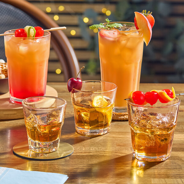 A clear glass tumbler filled with an amber beverage and ice, shown among other assorted drinks on a wooden table.