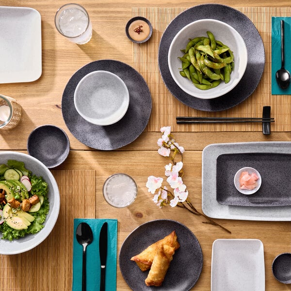 A light gray round melamine bowl placed on a dark plate, part of a table setting with various dishes and utensils.
