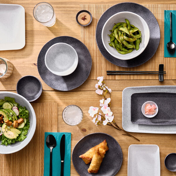 A large white round melamine bowl filled with edamame, placed on a dark plate as part of a table setting.