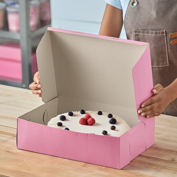 A pink quarter sheet cake box with a cake inside, being held open on a wooden counter.