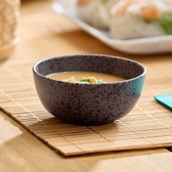 A small dark gray round melamine ramekin filled with soup, placed on a bamboo mat.