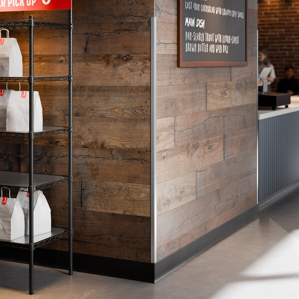 A stainless steel wall corner guard with adhesive strips installed on a wooden wall corner in a commercial setting.