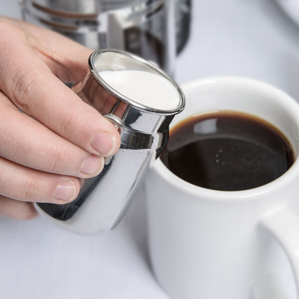 A hand pours American Metalcraft milk from a metal container into a cup of coffee.
