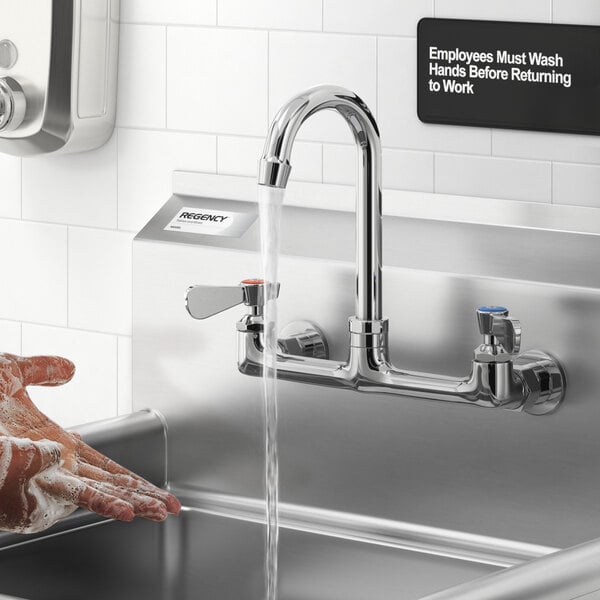 A person is washing their soapy hands under a wall-mounted, chrome gooseneck faucet in a commercial sink area.
