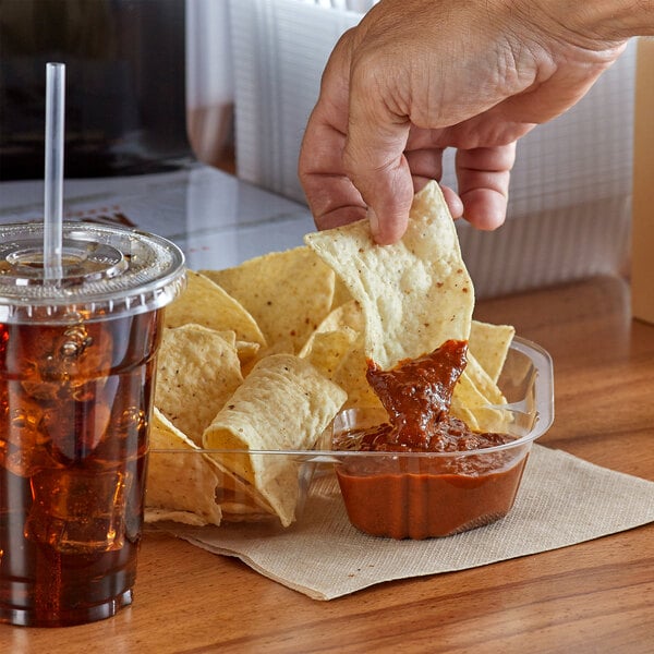 A hand dipping a tortilla chip into a bowl of chili sauce on a table with a drink and bowl of chips.