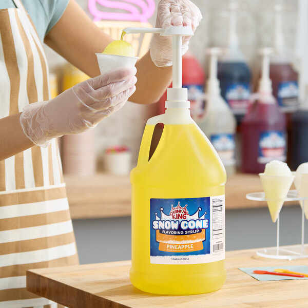 A gallon jug of Carnival King Pineapple Snow Cone Syrup with a pump dispenser being used to flavor a snow cone.
