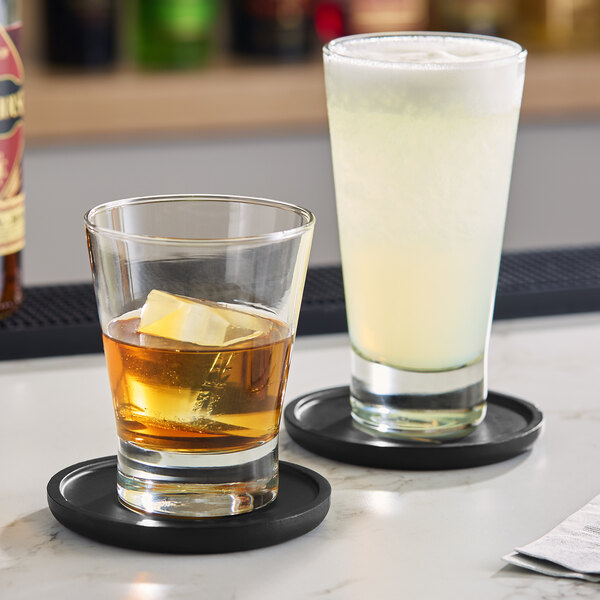 A clear Acopa Fusion 15.25 oz. beverage glass filled with a light-colored drink, shown next to a smaller glass with an amber liquid and ice, both on black coasters.