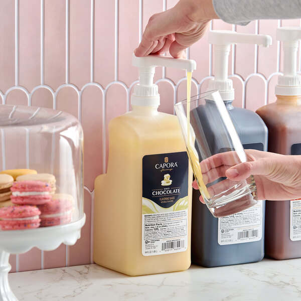 A large pump bottle of Capora White Chocolate Flavoring Sauce being dispensed into a glass.