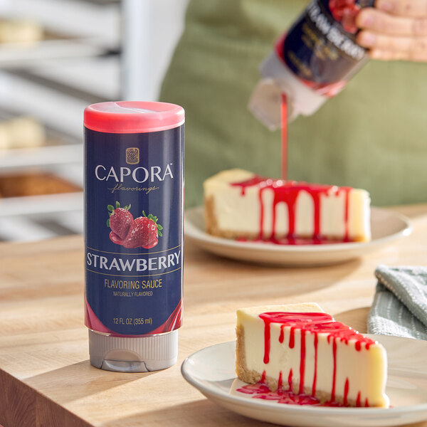 A 12 fl. oz. bottle of Capora Strawberry Flavoring Sauce placed on a wooden surface next to cheesecake slices drizzled with the sauce.