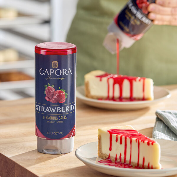 A bottle of Capora Strawberry Flavoring Sauce is shown on a wooden surface next to a slice of cheesecake topped with strawberry sauce.