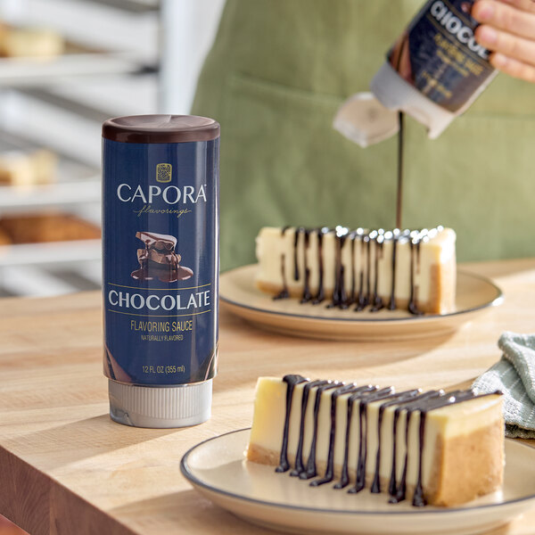 A bottle of Capora chocolate flavoring sauce is placed on a table next to plates of cheesecake drizzled with chocolate sauce, with a person pouring sauce in the background.