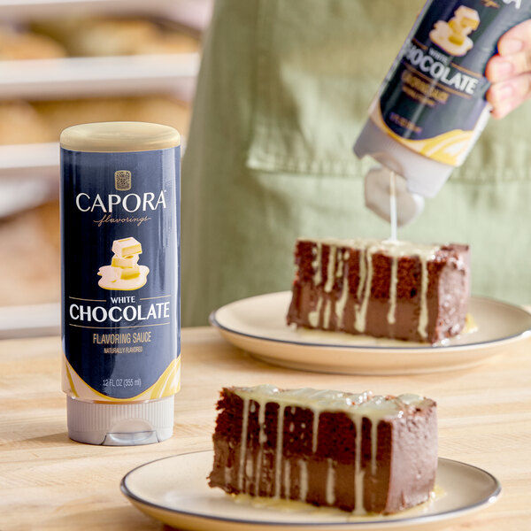 A bottle of Capora White Chocolate Flavoring Sauce, 12 oz, shown next to slices of chocolate cake being drizzled with the sauce.
