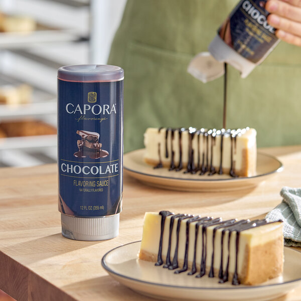 A 12 fl. oz. bottle of Capora Chocolate Flavoring Sauce placed on a table next to cheesecake slices drizzled with chocolate sauce.