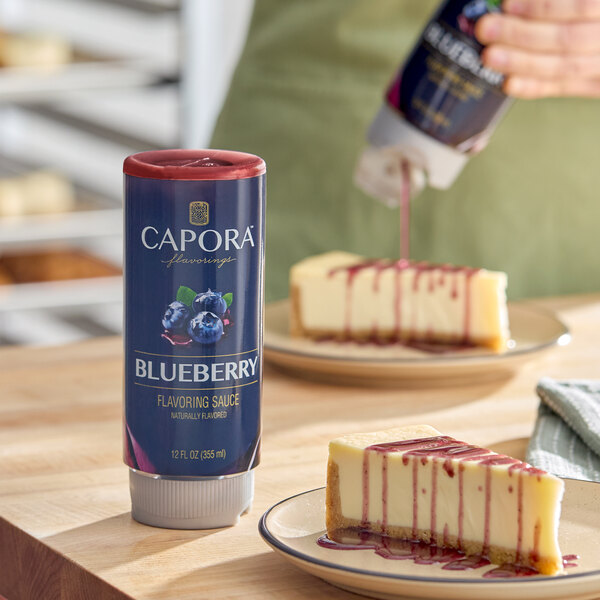 A 12 fl. oz. bottle of Capora Blueberry Flavoring Sauce displayed on a table next to cheesecake slices drizzled with the sauce.