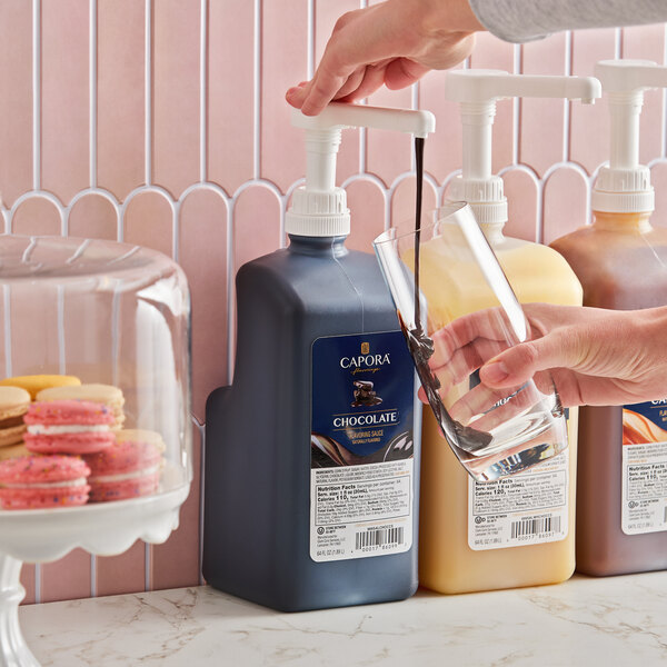A large bottle of Capora chocolate flavoring sauce with a pump dispenser being used to pour sauce into a glass.