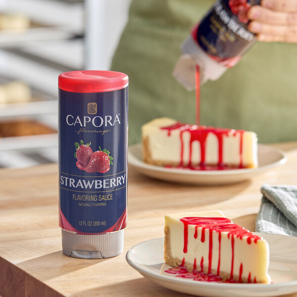 A 12 fl. oz. bottle of Capora Strawberry Flavoring Sauce displayed on a table next to cheesecake slices topped with the sauce.