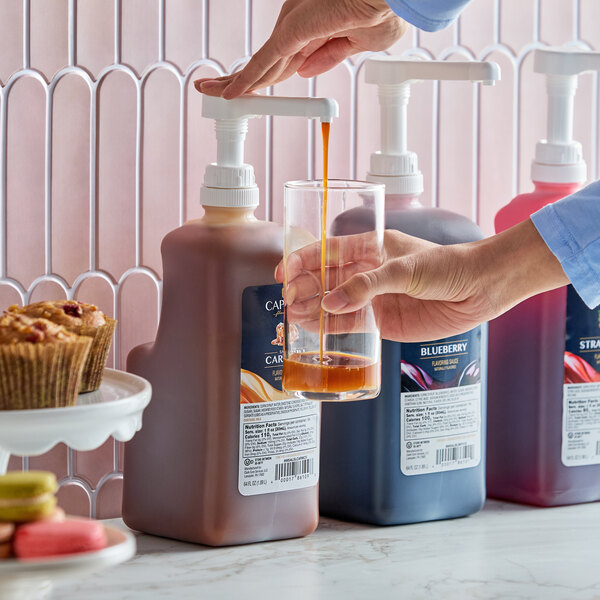 A large pump bottle of Capora Salted Caramel Flavoring Sauce being dispensed into a glass, with other flavoring sauces in the background.
