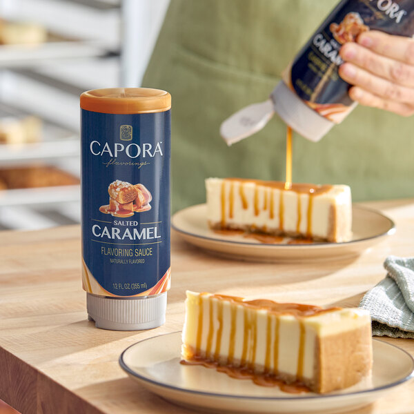 A bottle of Capora Salted Caramel Flavoring Sauce placed on a table next to slices of cheesecake drizzled with caramel sauce.