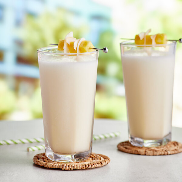 A pair of tall glasses filled with a creamy, light-colored pina colada smoothie, garnished with pineapple and coconut pieces on top.