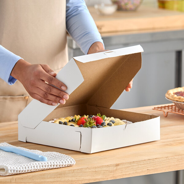 A white, square bakery box being opened to reveal a fruit-topped pie inside.