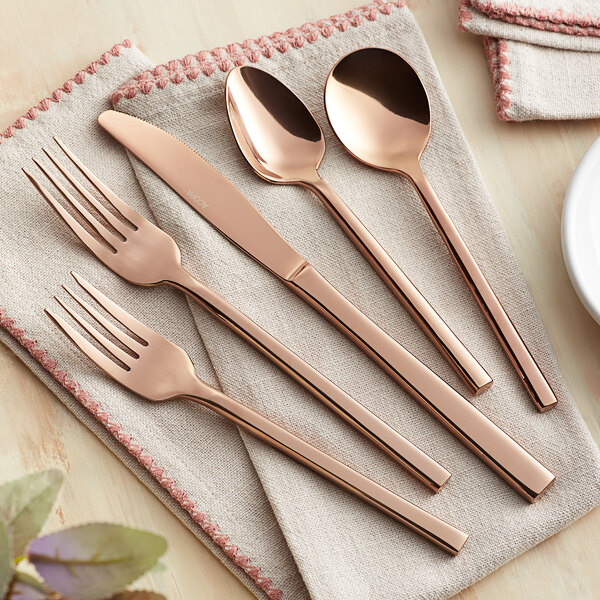 A five-piece set of rose gold stainless steel flatware including two forks, a knife, and two spoons, displayed on a beige napkin.