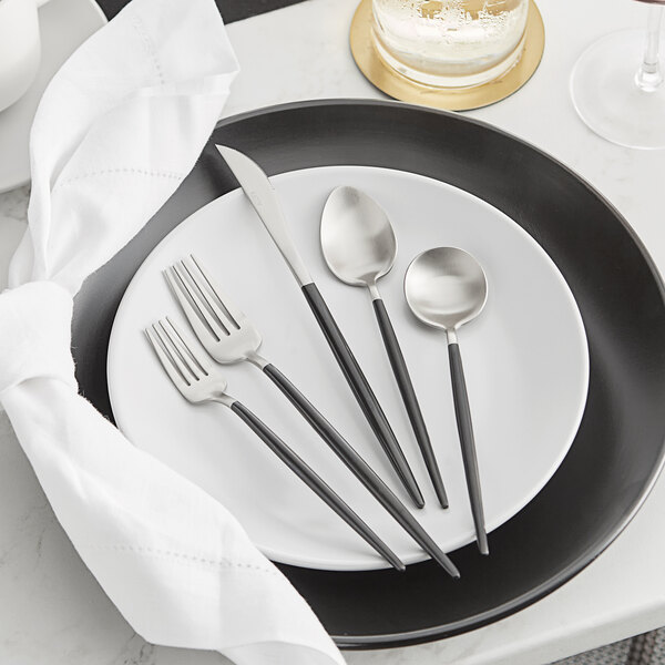 A five-piece set of black and silver brushed stainless steel flatware, including two forks, a knife, and two spoons, displayed on a white plate.