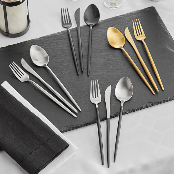A set of modern flatware in gold, black, and silver finishes displayed on a slate serving board.