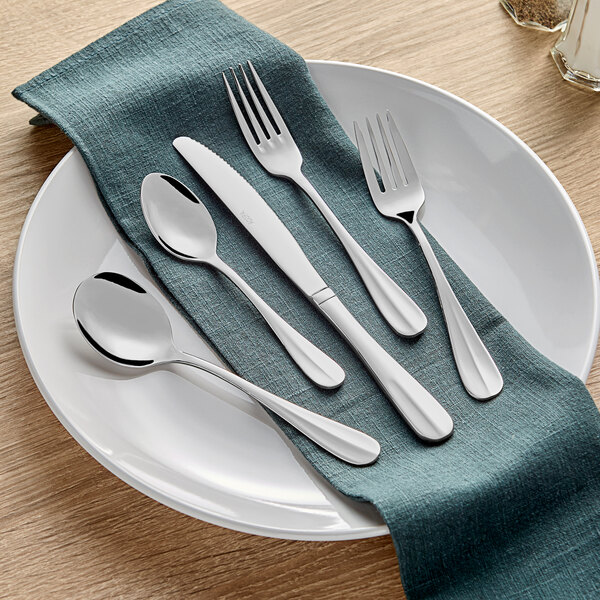 A five-piece set of stainless steel flatware including two forks, two spoons, and a knife, displayed on a white plate with a teal napkin.