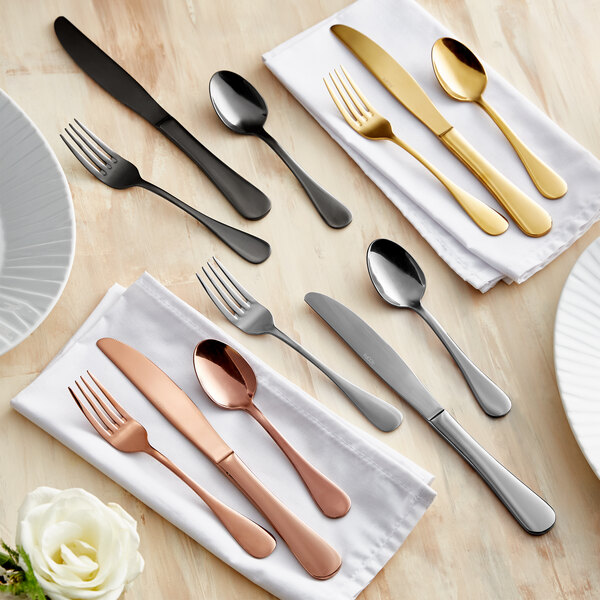 A set of five-piece stainless steel flatware in various finishes, including black, gold, silver, and copper, displayed on napkins.