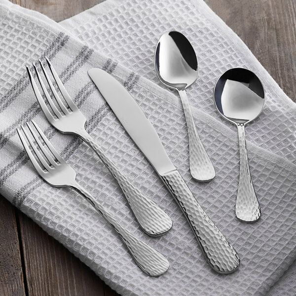 A five-piece set of stainless steel flatware with a hammered handle design, including two forks, a knife, and two spoons, displayed on a white textured cloth.