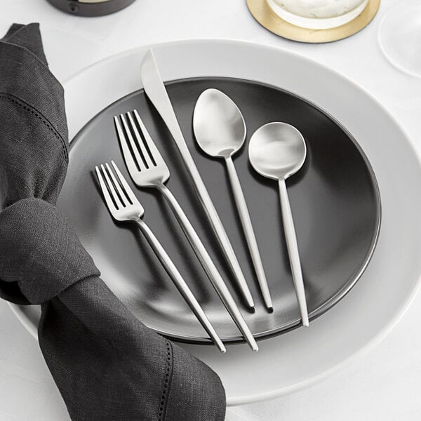 A five-piece set of brushed stainless steel flatware, including two forks, a knife, and two spoons, displayed on a black plate with a white plate and black napkin.
