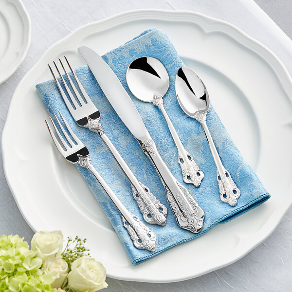 A five-piece set of ornate stainless steel flatware, including two forks, a knife, and two spoons, displayed on a blue napkin atop a white plate.