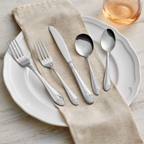 A five-piece set of stainless steel flatware with decorative handles, displayed on a white plate with a beige napkin.
